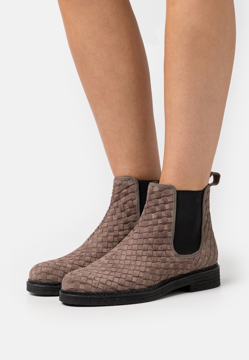 Bottines Chelsea en daim tissé marron avec des panneaux élastiques sur les côtés, bout rond et semelle extérieure en caoutchouc noir. Elles présentent une languette au niveau du talon.