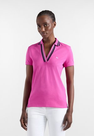 Nainen, joka käyttää kirkkaanpunaista lyhythihaista poolopaitaa, jossa on laivastonsininen ja valkoraidallinen kaulus, yhdistettynä valkoisiin housuihin, seisoo valkoista taustaa vasten.