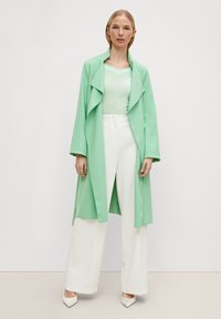 Cappotto lungo verde chiaro con colletto drappeggiato, abbinato a una maglietta leggera e pantaloni bianchi a gamba larga. Tessuto morbido e vestibilità rilassata.