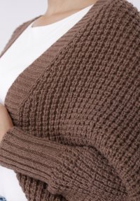 Καφέ πλεκτή ζακέτα με υφή μοτίβου, με ριμπ μανσέτες και ανοιχτό μπροστινό μέρος, φορεμένη πάνω από λευκό πουκάμισο.