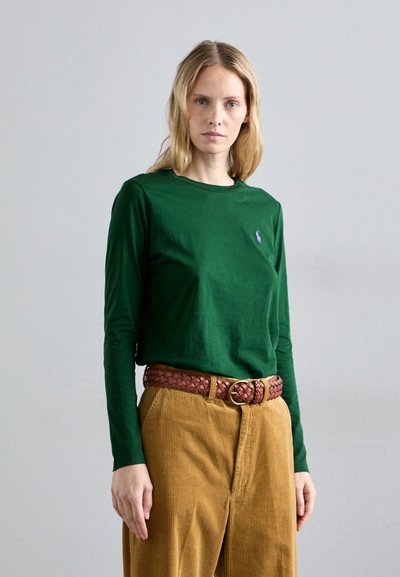 Donna con lunghi capelli biondi che indossa una maglietta verde a maniche lunghe, una cintura intrecciata marrone e pantaloni in corduroy senape su uno sfondo neutro.