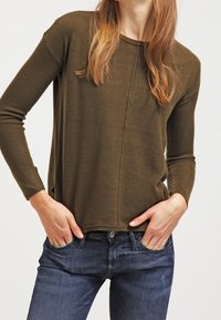 Suéter ligero de punto en verde oliva, con mangas largas y cuello redondo. Presenta una textura acanalada y un corte relajado. Llevado con jeans oscuros.