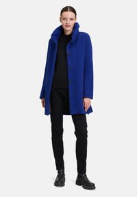 Manteau en laine bleu avec un col haut, fermeture à boutons et une finition texturée. Le modèle porte un haut noir et un pantalon avec des bottines.
