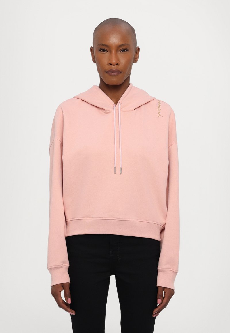 Sweatshirt rosa com capuz e design cropped, com cordão e punhos canelados. Detalhe em dourado notável no ombro. Usado com calças pretas.