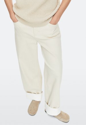 Personne portant un pantalon crème à jambes larges avec de grands revers, un pull en maille beige et des chaussures en daim marron clair sans lacets, debout les mains dans les poches.