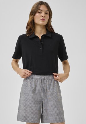 Kvinde justerer sort kortærmet polo skjorte, der er stoppet ned i højtaljede grå og hvide stribede shorts med snøre, på ensartet baggrund.