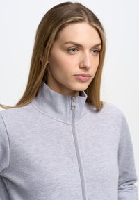 Svijetlosiva jakna na zip s visokim ovratnikom, rebrastom teksturom i glatkom površinom s suptilnim heather uzorkom.