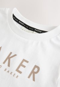 T-shirt en coton blanc avec un col rond, une étiquette noire et un logo "BAKER" doré imprimé à l'avant. Texture lisse, design décontracté.