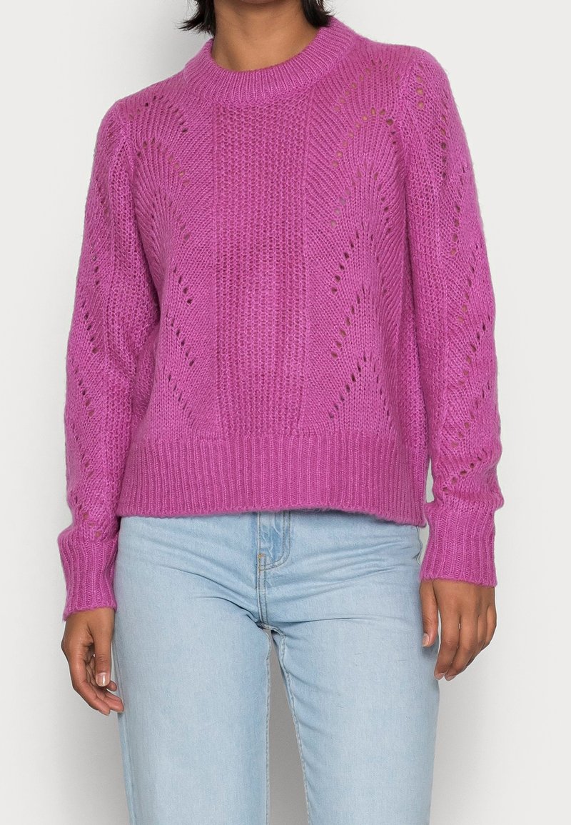 Rosa stickad tröja med en texturerad mönster och öppna sömdetaljer. Har ribbade ärmslut och nederkant, bärs över ljusblå jeans.