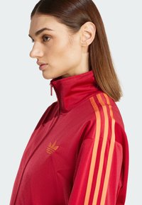 Rote sportliche Zip-Jacke mit hohem Kragen, ausgestattet mit orangefarbenen drei Streifen-Akzenten auf den Schultern und einem kleinen Logo auf der linken Brust. Glatte Textur.