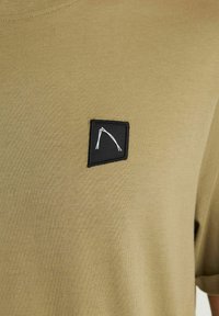 Alyvuogių spalvos medvilninis marškinėliai su trumpomis rankovėmis ir apvalia iškirpte. Papuoštas juodu kvadratiniu pleistru su baltu logotipu.