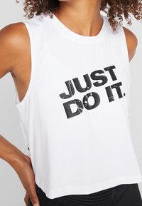 Donna che indossa un top bianco senza maniche con testo nero usurato "JUST DO IT." e mani appoggiate sui fianchi.