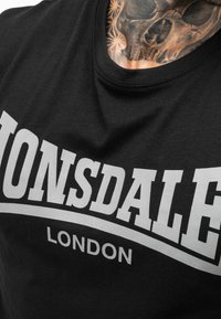 Camiseta negra de algodón con un prominente logo en gris "Lonsdale London" en letras negritas en el pecho. Textura suave, cuello redondo.