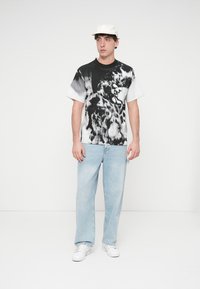 Schwarzes und weißes Grafik-T-Shirt mit abstraktem Design, kombiniert mit hellblauen Denim-Jeans und weißen Sneakers. Weiße Kappe getragen.