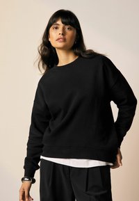 Zwarte sweatshirt van zacht materiaal, met een ronde hals, lange mouwen en geribbelde boorden en zoom. Gelaagd over een witte top.