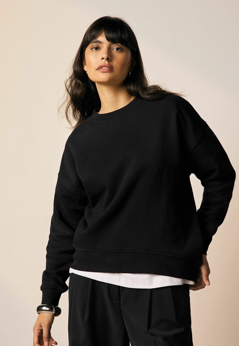 Zwarte sweatshirt van zacht materiaal, met een ronde hals, lange mouwen en geribbelde boorden en zoom. Gelaagd over een witte top.