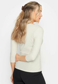 Hellbeige, geripptes Langarmshirt mit eng anliegendem Design. Verfügt über einen runden Ausschnitt und ist an den Seiten geöffnet, getragen mit schwarzen Hosen.