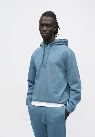 Sweat à capuche en coton bleu avec cordon de serrage et poche avant, assorti à un pantalon de survêtement. Texture lisse, coupe décontractée, détail de marque minimaliste.