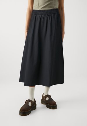 Person wearing a high-waisted black midi skirt, light socks, brown chunky shoes, and a sleeveless light green top, standing indoors.