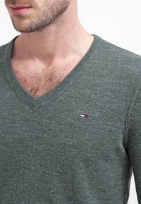 Zielony sweter w serek wykonany z miękkiego materiału, z małym haftowanym logo w czerwono-biało-niebieskich akcentach na piersi.