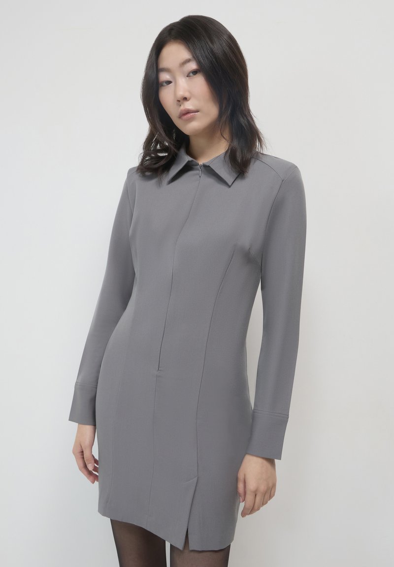 Abito grigio a maniche lunghe con colletto, zip frontale e spacchi laterali. Realizzato in tessuto liscio con una silhouette aderente.