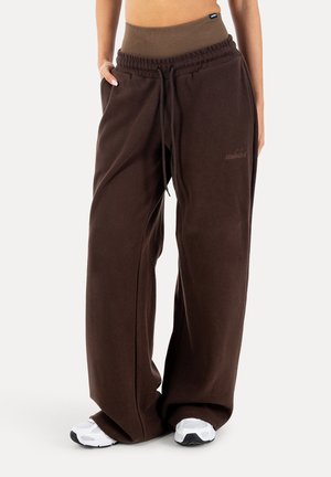 Kvinde iført løse brune joggingbukser med elastisk taljebånd og snore, kombineret med hvide sneakers, stående mod en hvid baggrund.