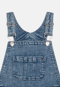 Pantalones de mezclilla con tirantes ajustables de metal, lavado azul y un bolsillo frontal con una pequeña etiqueta de logo. Tejido texturizado con costuras visibles.