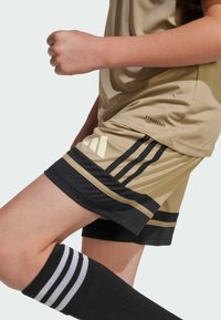 Jersey y pantalones cortos deportivos beige con acentos negros, que presentan tres rayas negras en los pantalones cortos, logo AEROREADY y calcetas negras hasta la rodilla con rayas blancas.
