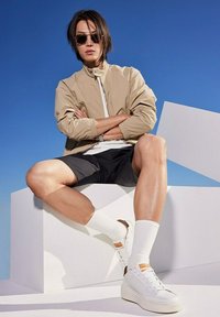 Beige Bomberjacke, weißes T-Shirt, schwarze Shorts, weiße Socken, weiße Turnschuhe mit braunen Akzenten; im Sitzen vor einem geometrischen weißen Hintergrund.