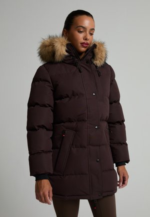 Femme portant un manteau d'hiver matelassé marron foncé avec une capuche bordée de fourrure, debout devant un fond gris clair uni.