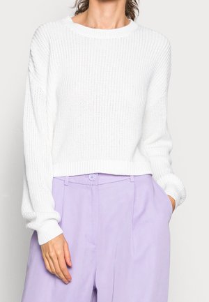 Kobieta w białym dzianinowym swetrze z długim rękawem, włożonym do wysokowzsitowych lawendowych spodni, z jedną ręką w kieszeni, a drugą swobodnie opadającą.