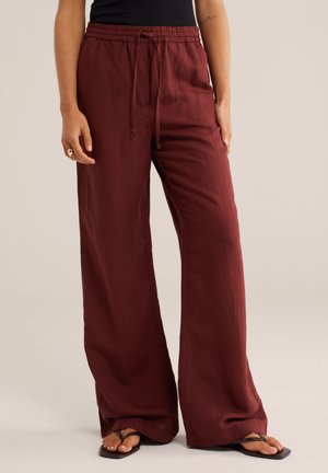 Vrouw draagt losse, wijd uitlopende bordeauxrode broek met trekkoord in de taille, zwart mouwloos shirt, ring aan linkerhand en zwarte sandalen.