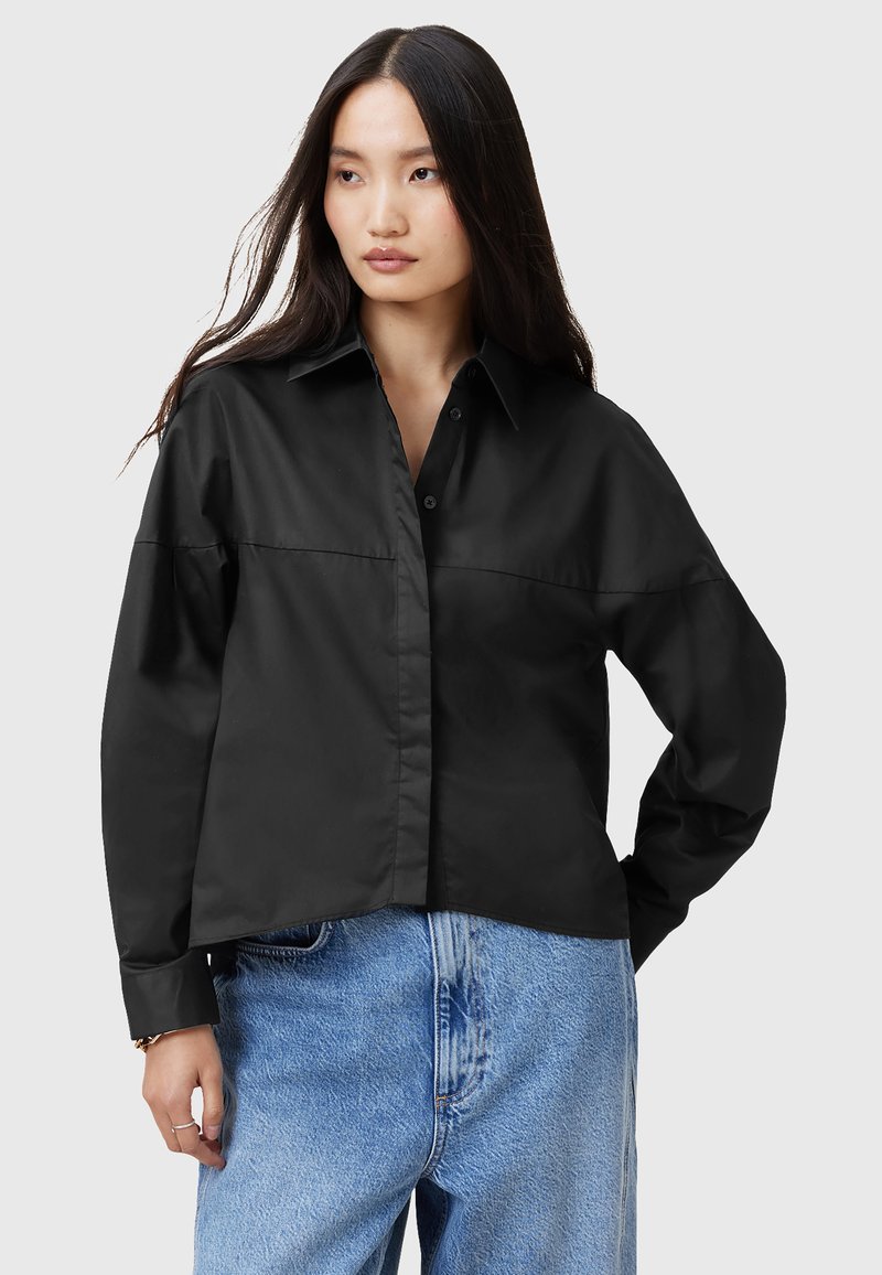 Camicia nera con colletto aperto, maniche lunghe e vestibilità ampia, realizzata in un tessuto liscio. Abbinata a jeans in denim blu chiaro.