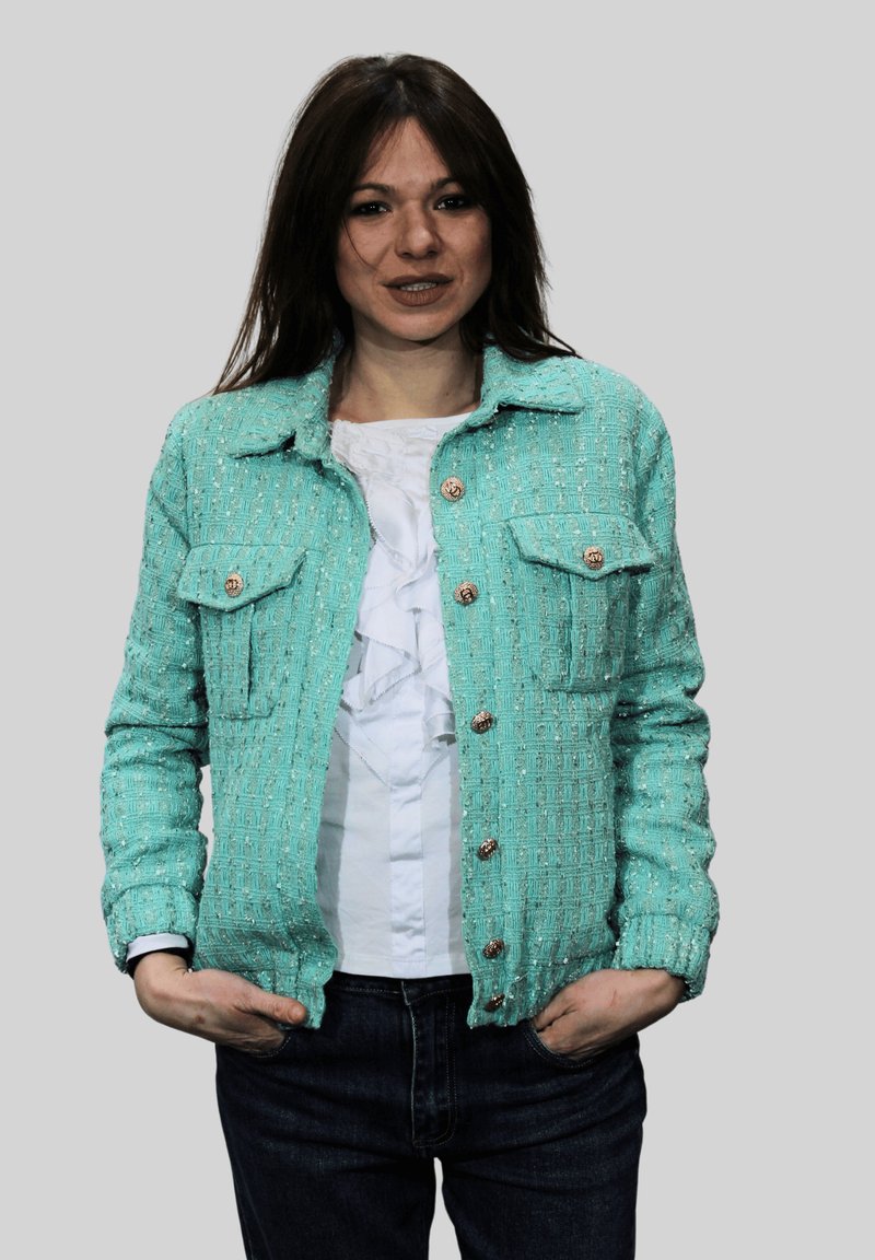 Chaqueta de tweed verde azulado claro con dos bolsillos en el pecho, botones dorados y un patrón texturizado. Llevada sobre un top blanco con volantes y jeans oscuros.