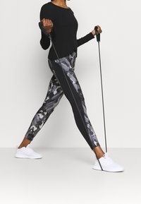 Czarna bluzka z długim rękawem w połączeniu z szarymi legginsami we wzory; legginsy mają boczny pasek. Białe buty sportowe i bandaże oporowe trzymane w dłoniach.