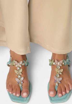 Pies con sandalias turquesas adornadas con mariposas de cristal multicolores, combinadas con pantalones beige de pierna ancha.