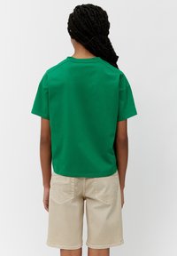 Una persona con el cabello trenzado lleva una camiseta verde de manga corta y pantalones cortos beige hasta la rodilla, de espaldas, frente a un fondo blanco liso.