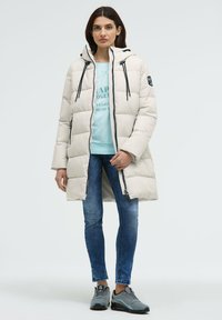 Puffermantel in hellbeige mit Kapuze, ausgestattet mit schwarzen Kordeln und einem Reißverschluss. Kombiniert mit einem blauen Sweatshirt, engen Jeans und grauen Sneakers.