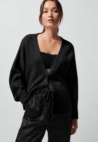 Schwarzer gerippter Cardigan mit tiefem V-Ausschnitt, Knopfverschluss und übergroßen Ärmeln, kombiniert mit einem engen schwarzen Oberteil und schwarzen Satin-Hosen.