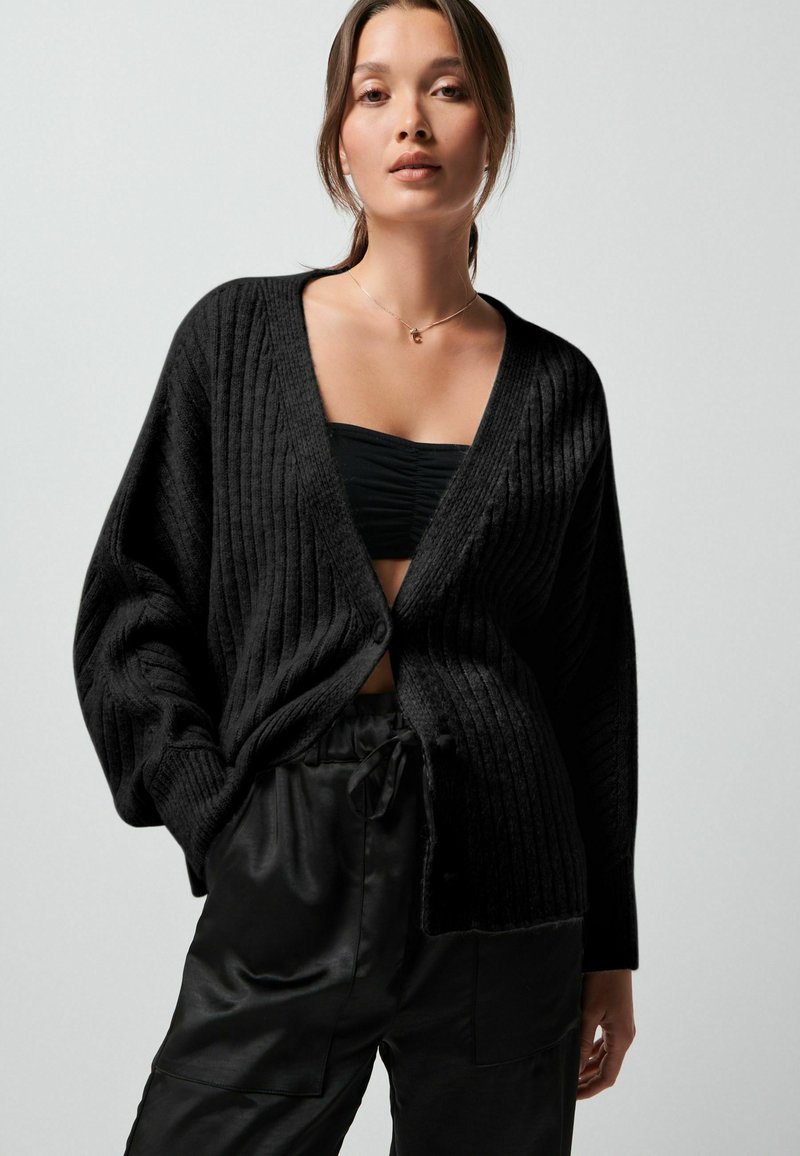 Schwarzer gerippter Cardigan mit tiefem V-Ausschnitt, Knopfverschluss und übergroßen Ärmeln, kombiniert mit einem engen schwarzen Oberteil und schwarzen Satin-Hosen.