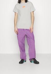 Person wearing a light gray Nike t-shirt with orange logo, loose purple pants, and black sneakers with teal and orange accents, standing on white floor.