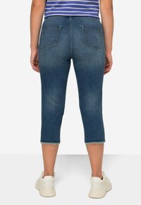Pantalones capri de mezclilla en azul oscuro con un acabado desgastado, que cuentan con dos bolsillos traseros y dobladillos deshilachados. Se llevan con zapatillas blancas.