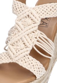 Sandalia de yute beige con correas entrelazadas hechas de cuerda gruesa, que presenta un acento trenzado y una plantilla plana de textura marrón.