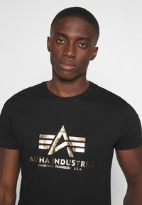 Fekete pamut póló fémes arany logóval, amely háromszög alakú és az "ALPHA INDUSTRIES" felirat található a mellrészen. Rövid ujjú.