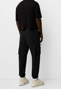 Svart bomulls t-shirt med kort ärm i kombination med svarta cargo-byxor med sidofickor och elastiska manschetter, ihop med beigea träningsskor.