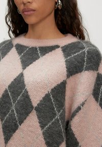 Vrouw in een pluizige roze en grijze ruitjestrui met zilveren oorringen, waarbij haar nek en schouders zichtbaar zijn.