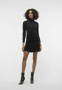 Un haut noir à manches longues associé à une mini-jupe noire ajustée. Le mannequin porte des bottines noires avec une finition texturée et un design fuselé.