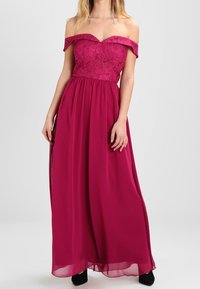 Off-the-shoulder klänning i fuchsia med spetsliv, flödande chiffongkjol och ankellång design. Har en åtsittande midja och mjuk textur.