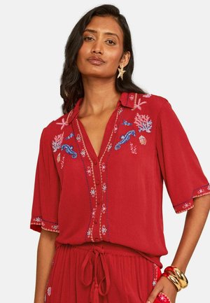 Vrouw draagt een rode blouse met geborduurde zeewezens en koraal, gecombineerd met een bijpassende rode broek en gouden en rode armbanden.