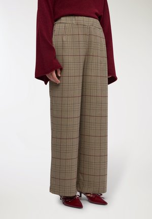 Pantaloni a gamba larga in tessuto beige a fantasia con quadri rossi e neri, completati da una vita alta e abbinati a scarpe color bordeaux.